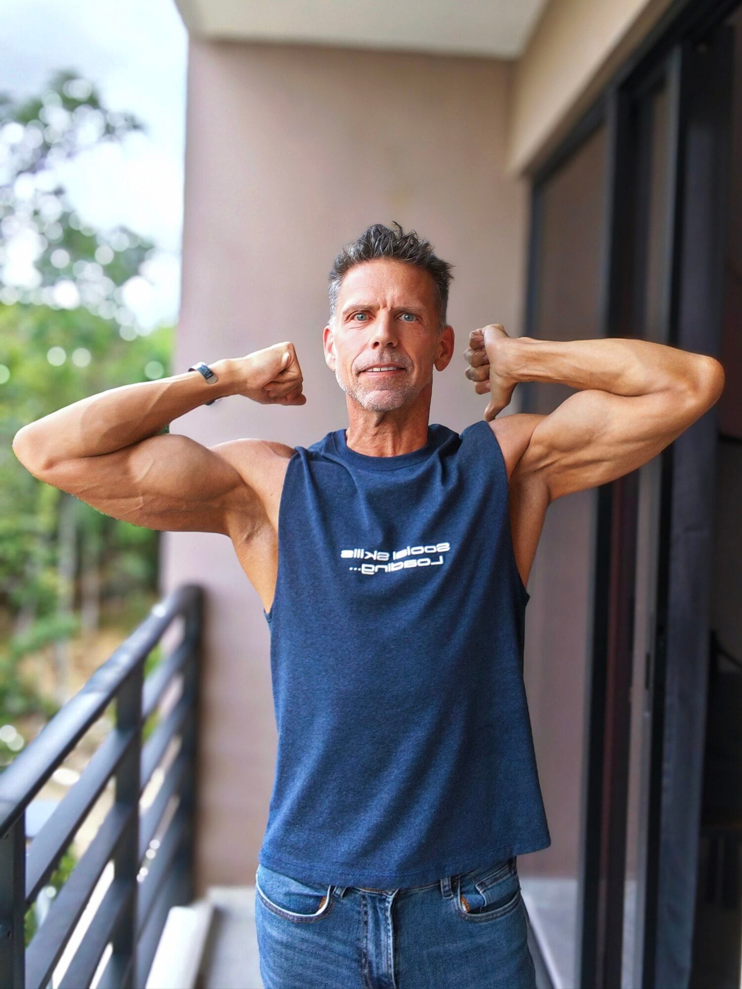 Eric standing on patio flexing and smiling with sleeveless blue shirt.