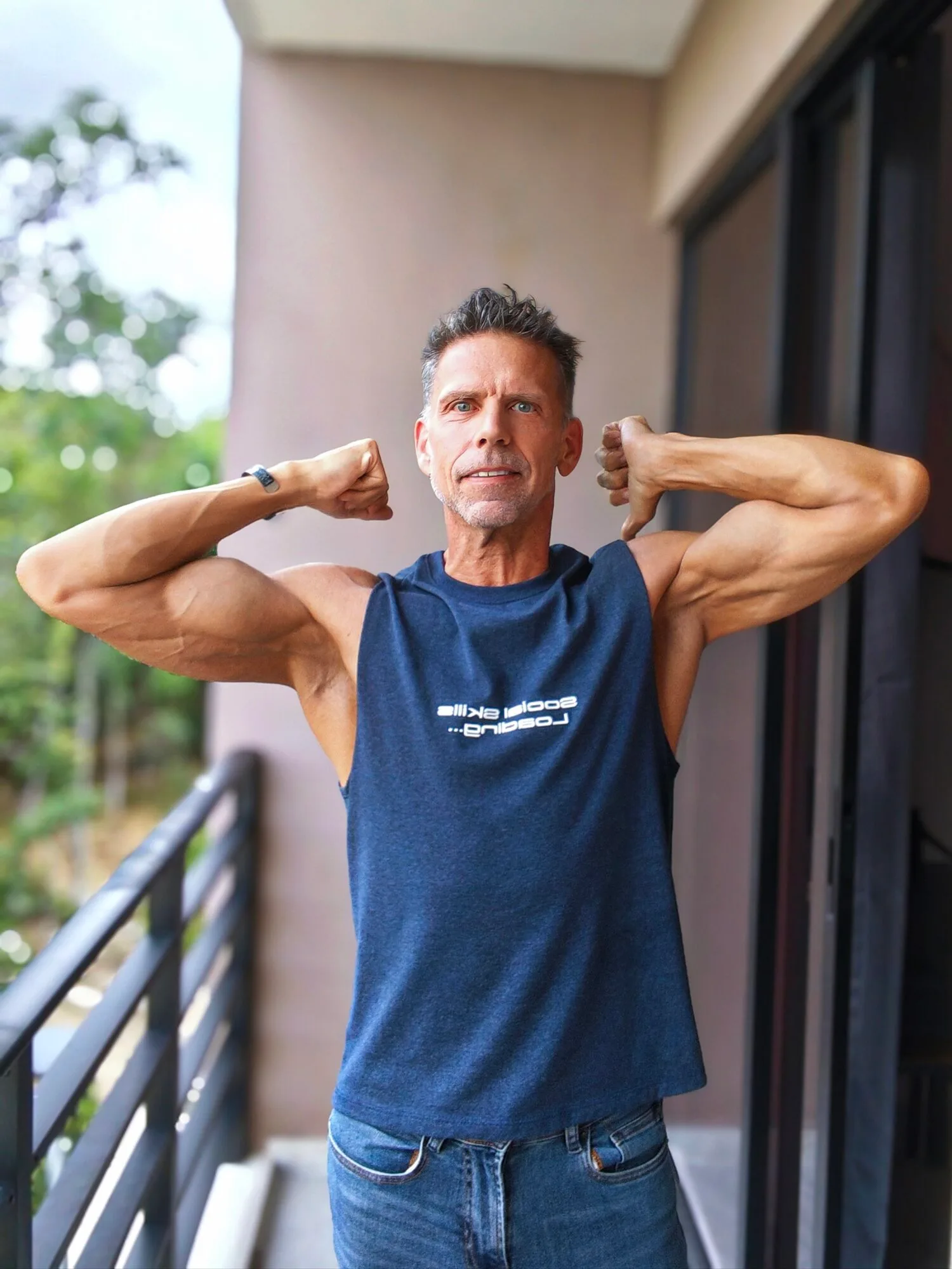 Eric standing on patio flexing and smiling with sleeveless blue shirt.